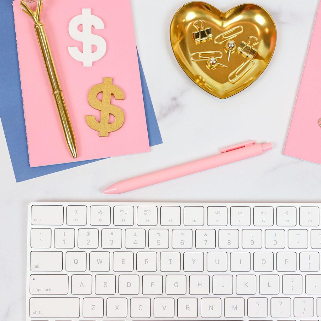 Picture of a keyboard with pink and blue papers, pens, a gold heart, and money signs