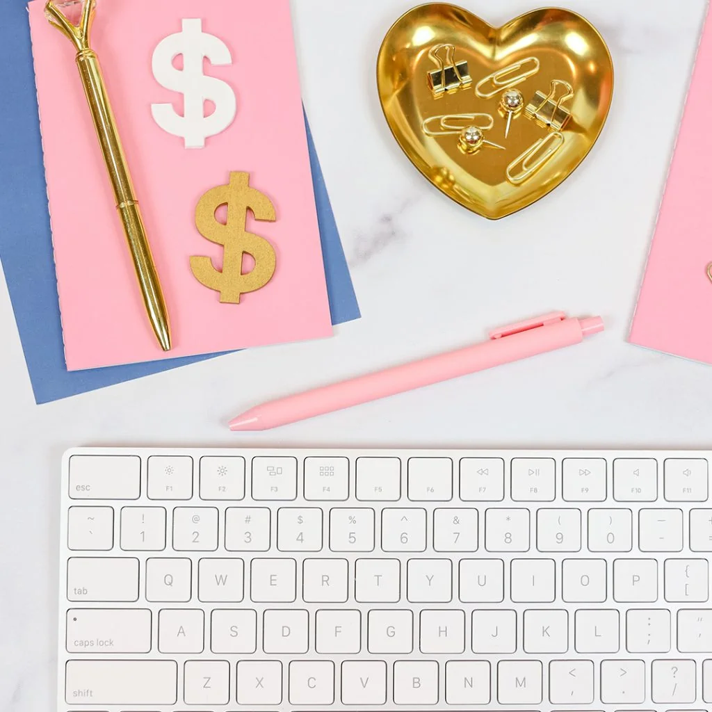 Picture of a keyboard with pink and blue papers, pens, a gold heart, and money signs
