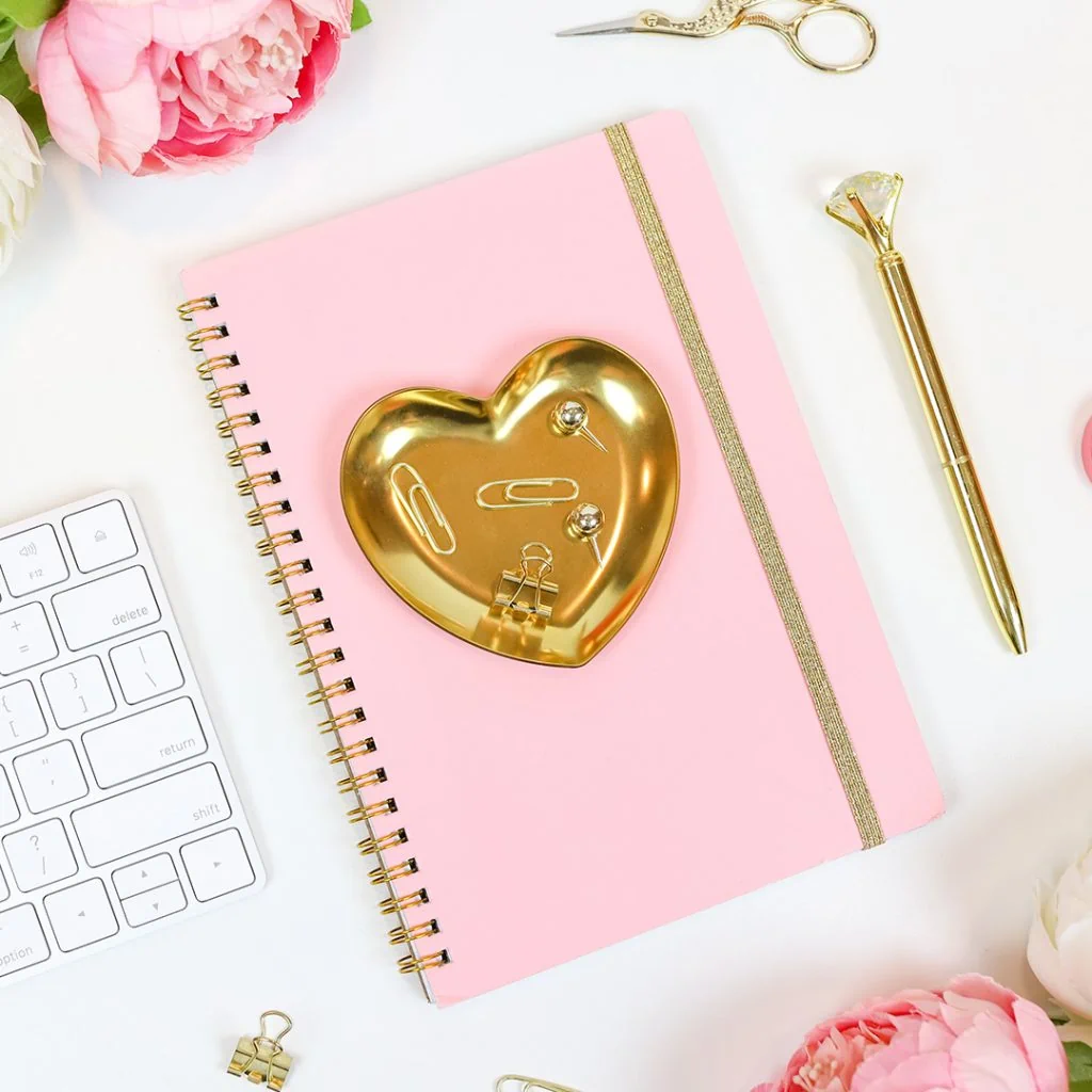 Picture of a gold heart on top of a light pink notebook with a keyboard and pen
